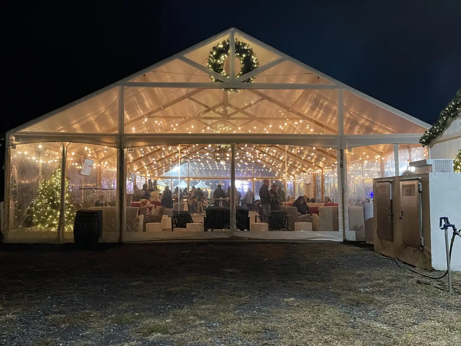 Guests inside a string lit clear tent with an evergreen wreath at an evening holiday party